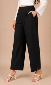 A model wearing black wide-leg cropped pants with side slits, paired with a white long-sleeve crop top and white heeled sandals, standing against a solid beige studio background.