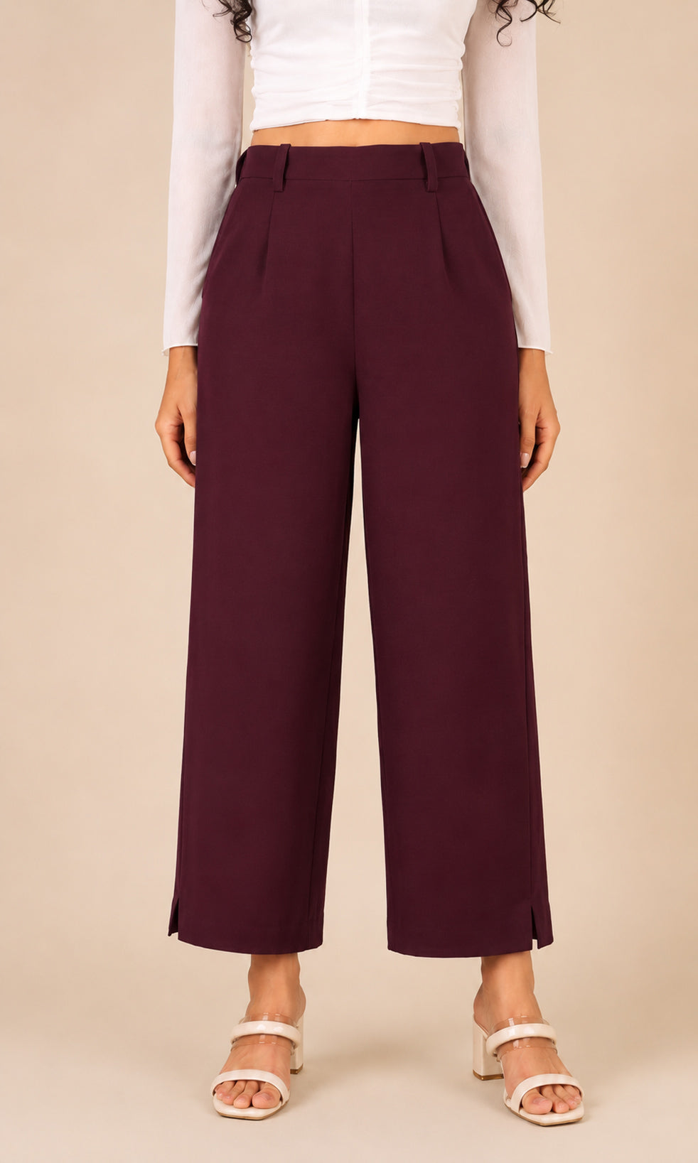 A model wearing mid-rise, wide-leg cropped pants in a deep burgundy color, paired with a white long-sleeve crop top and white heeled sandals, set against a solid beige background.