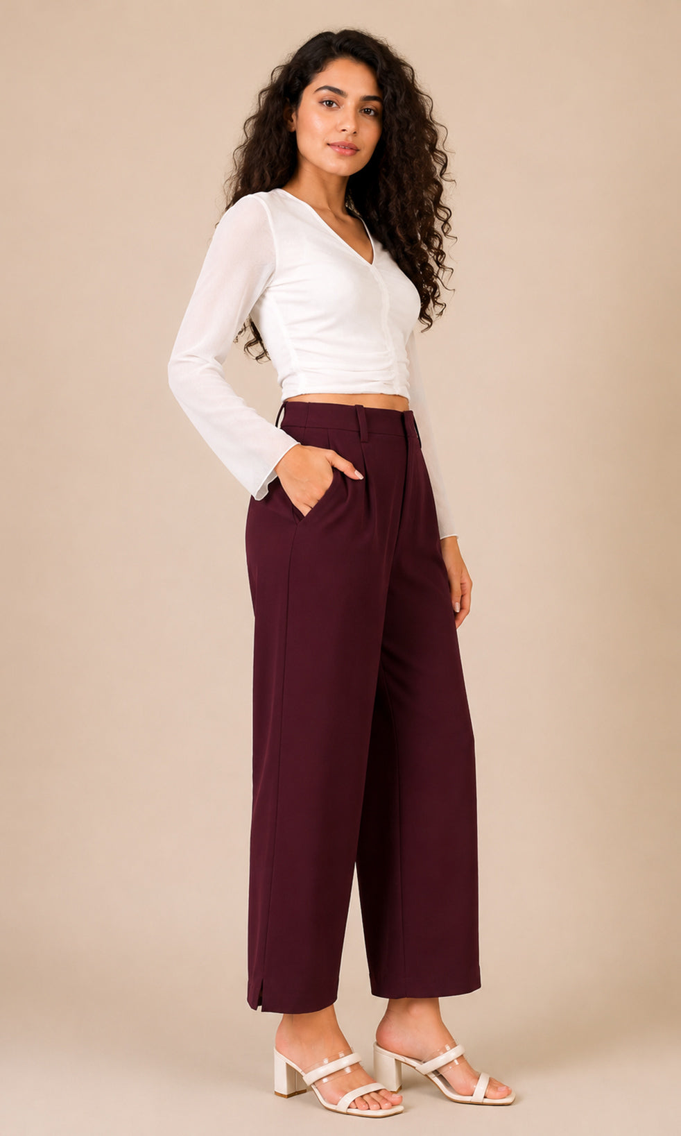 A woman modeling a white long-sleeve crop top and high-waisted burgundy wide-leg pants, paired with white heeled sandals, against a solid beige studio background.
