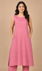 A woman modeling a sleeveless pink cotton kurta with side slits and matching pink trousers, standing against a solid beige studio background.