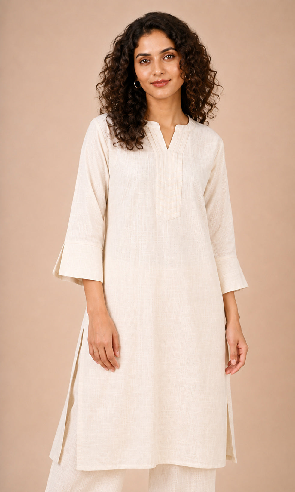 A woman with curly dark hair wearing a cream-colored cotton kurta with three-quarter sleeves and a split neckline, paired with matching cream trousers, set against a solid beige studio background.