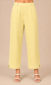A person wearing pale yellow cropped wide-leg pants with side slits and white heeled sandals, standing against a plain beige studio background.