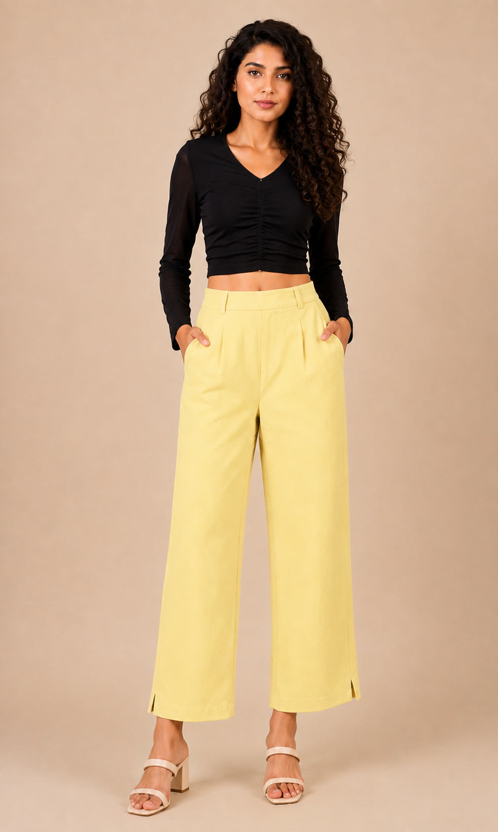 A woman modeling a pair of light yellow wide-leg cropped pants with a black long-sleeve ruched crop top, standing against a solid beige studio background.