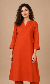 A woman modeling a rust-orange cotton kurta with three-quarter sleeves and a split neckline, paired with matching pants, set against a solid beige studio background.