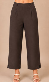 A person wearing dark brown cropped wide-leg pants with side slits and pleats, paired with white heeled sandals, against a plain beige studio background.
