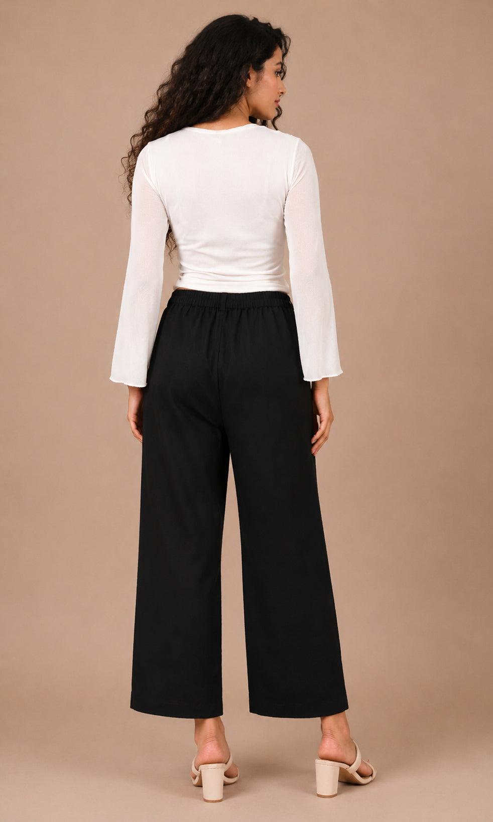 A woman shown from the back wearing a white long-sleeve top and black wide-leg cropped pants, standing against a plain beige studio background.