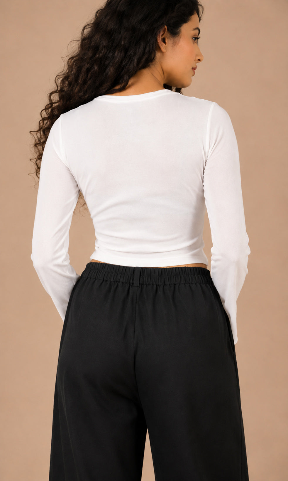 A woman shown from the back wearing a fitted white long-sleeve crop top and black wide-leg pants, standing against a plain warm beige studio background.