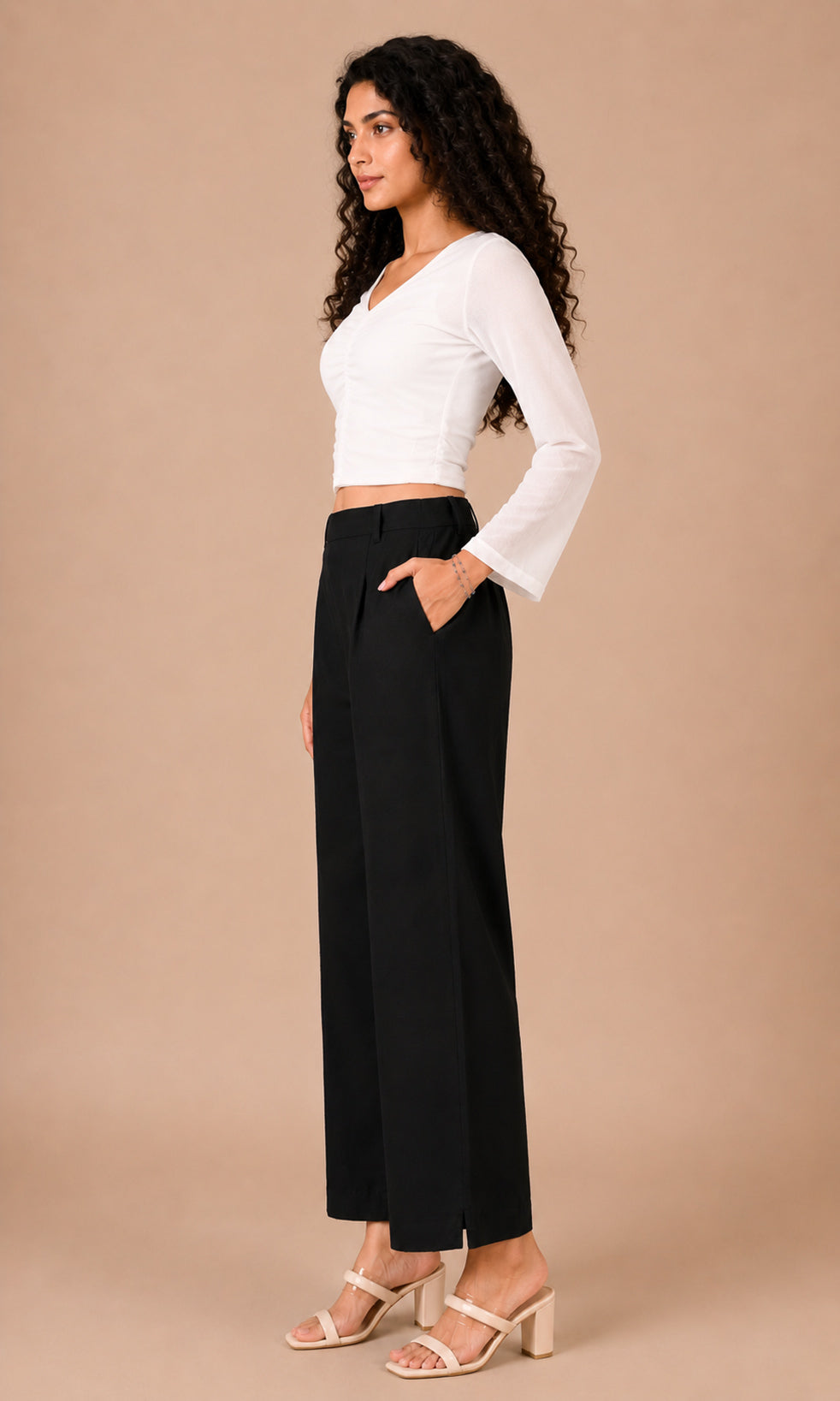 A woman modeling a white long-sleeve crop top and black wide-leg pants against a solid beige studio background.