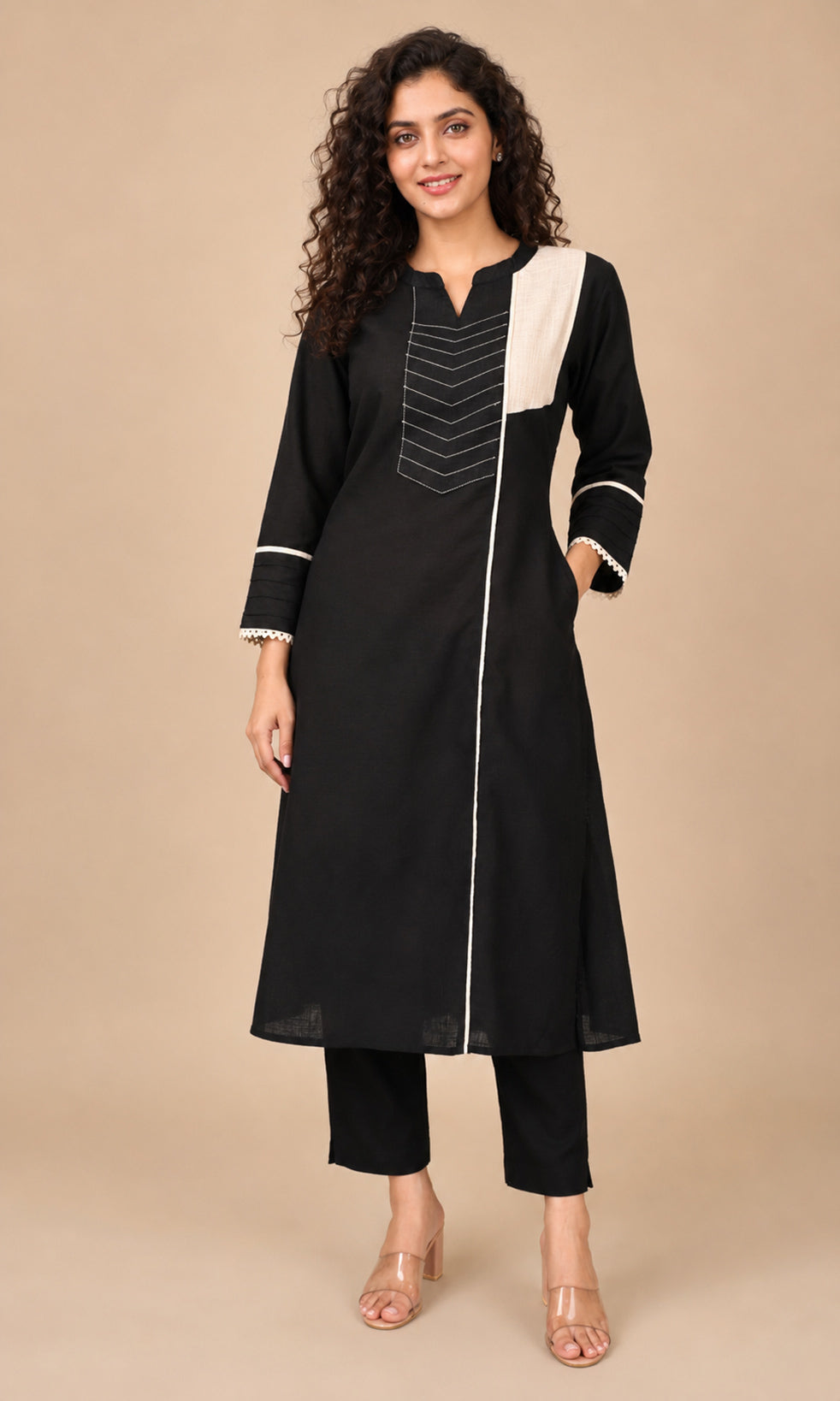A woman modeling a black long kurta with white contrast panels and white piping details, paired with black cropped pants and clear high-heeled sandals, against a solid beige studio background.