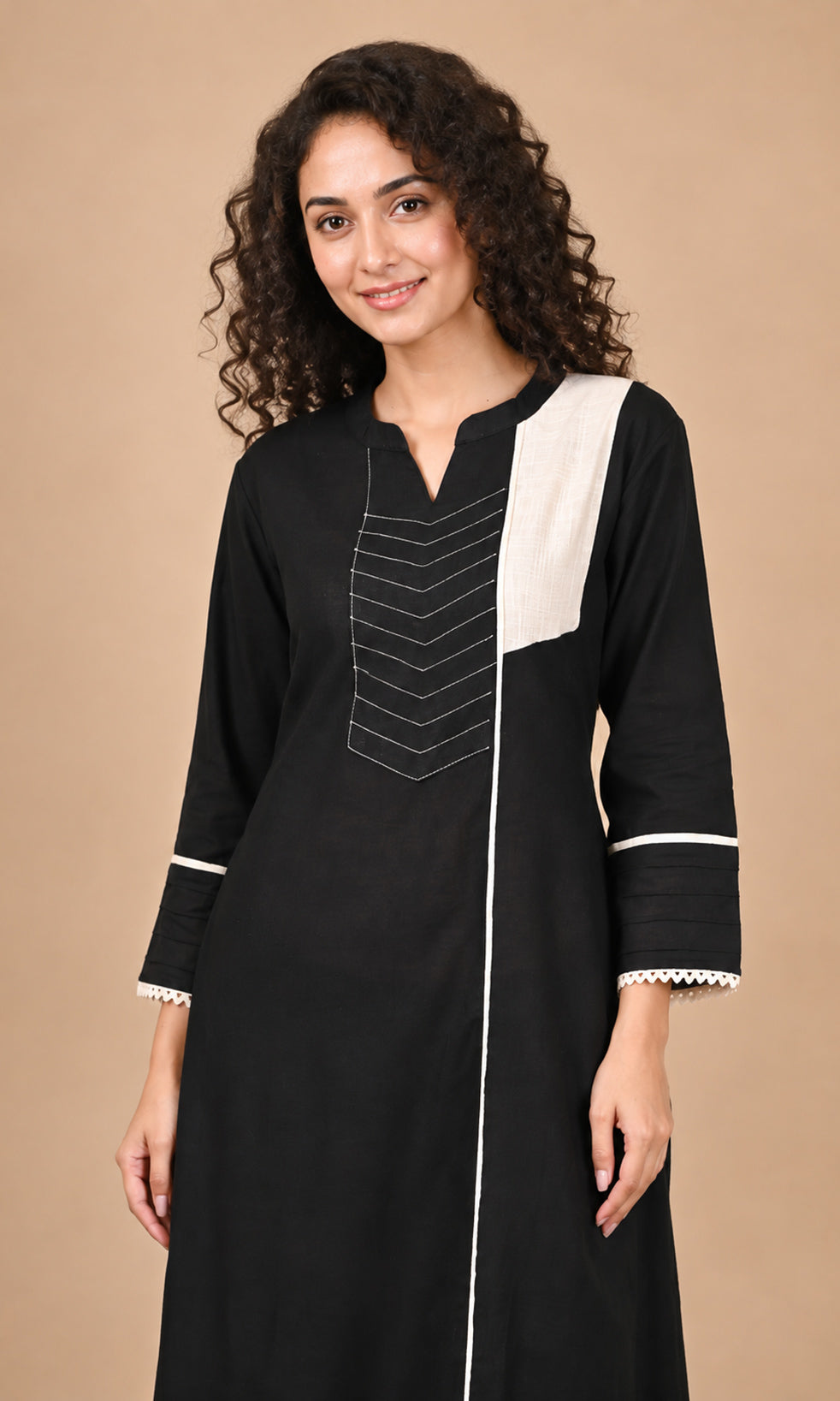 A woman modeling a black long kurta with white contrast panels on the chest and cuffs, featuring white geometric embroidery on the front, set against a solid beige background.