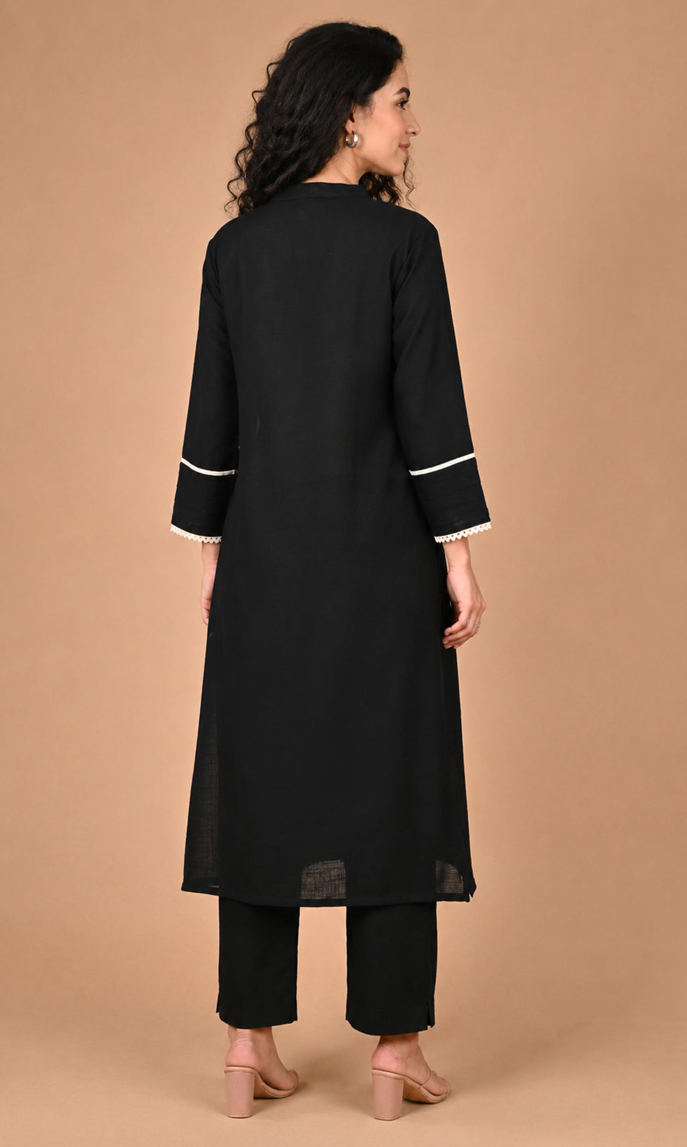 A woman shown from the back wearing a black long kurta with white contrast trim at the cuffs and black pants, standing against a solid tan studio background.
