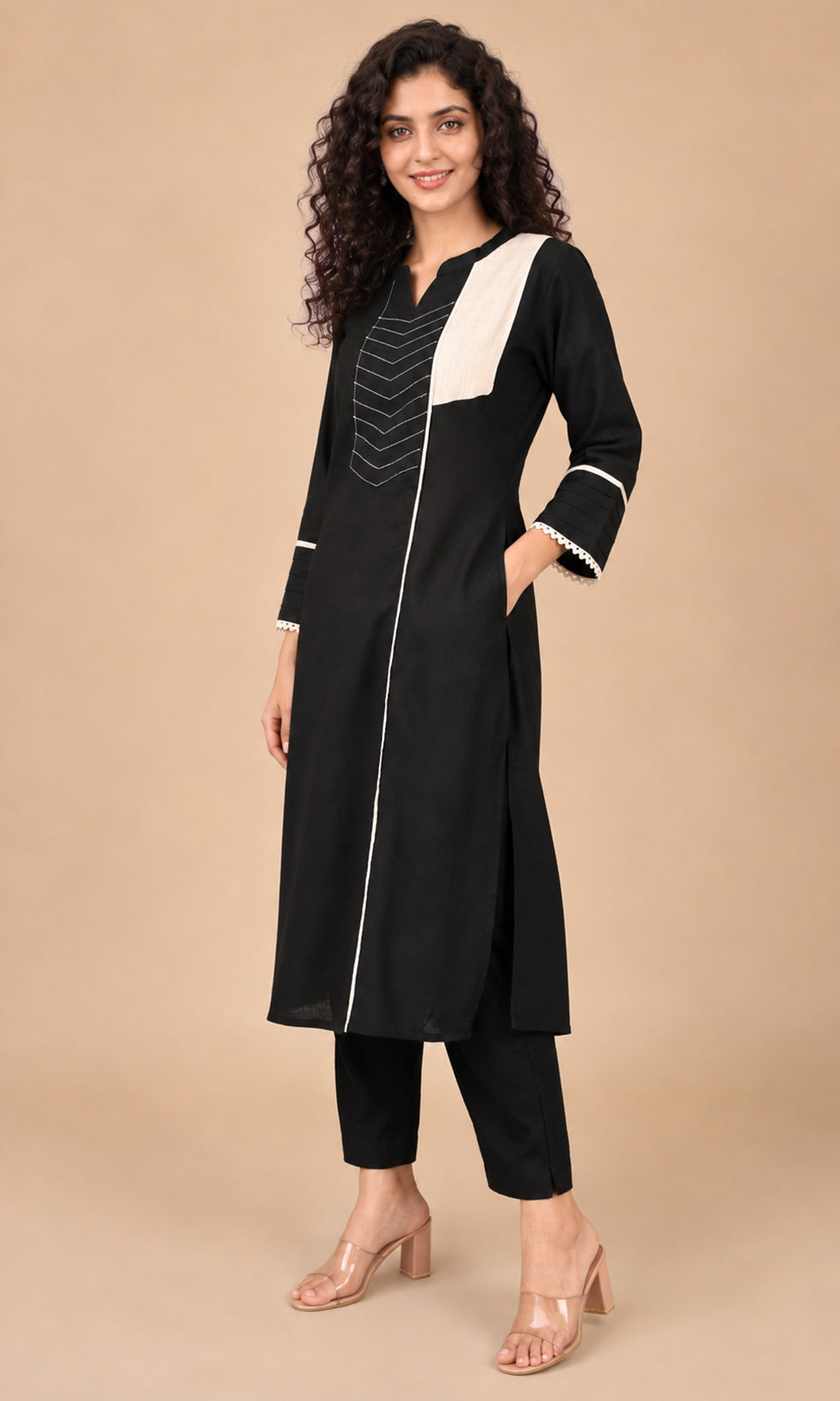 A woman modeling a black long kurta with contrasting white panels on the chest and cuffs, paired with black cropped pants and clear high-heeled sandals, against a solid beige studio background.