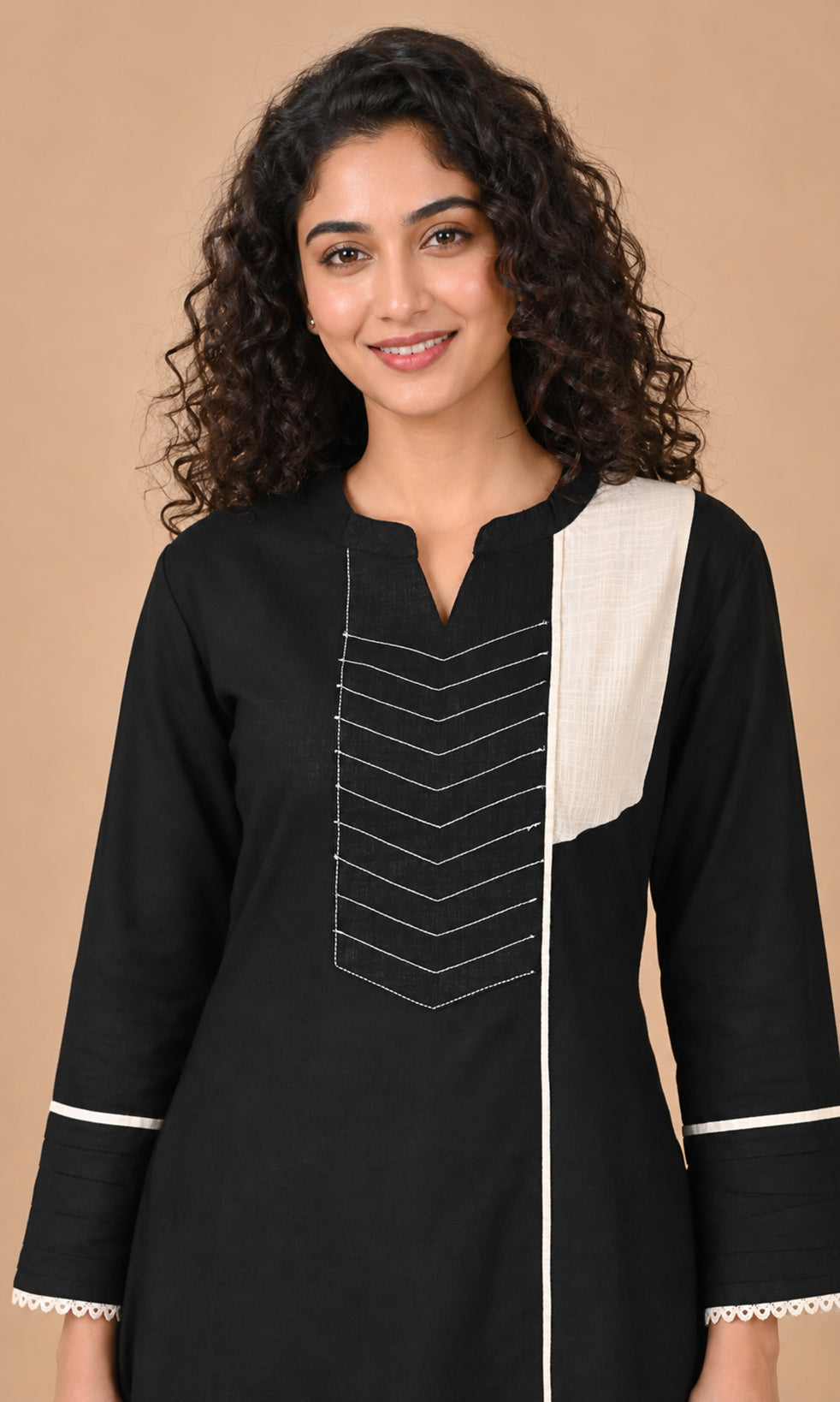 A woman wearing a black long-sleeve kurta with a contrasting off-white vertical panel on the right side, white chevron embroidery on the chest, and white piping on the cuffs, set against a solid beige background.