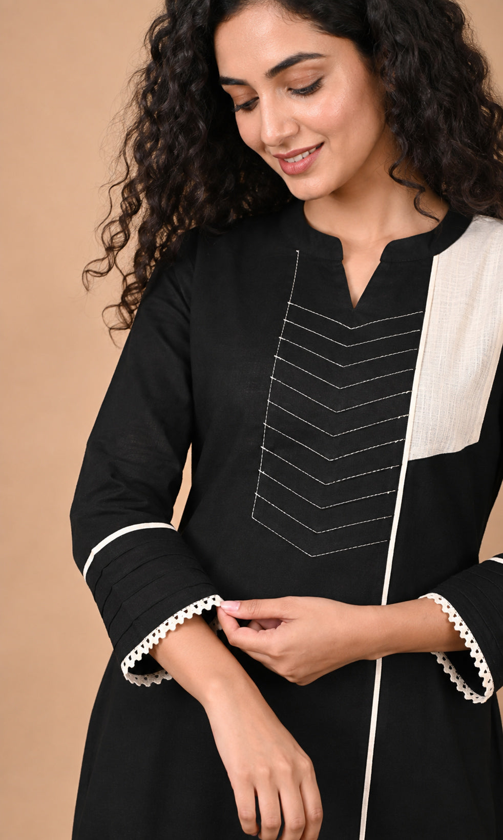 A woman wearing a black kurta with white contrast panels, white lace trim on the cuffs, and white geometric embroidery on the chest, posed against a solid beige background.