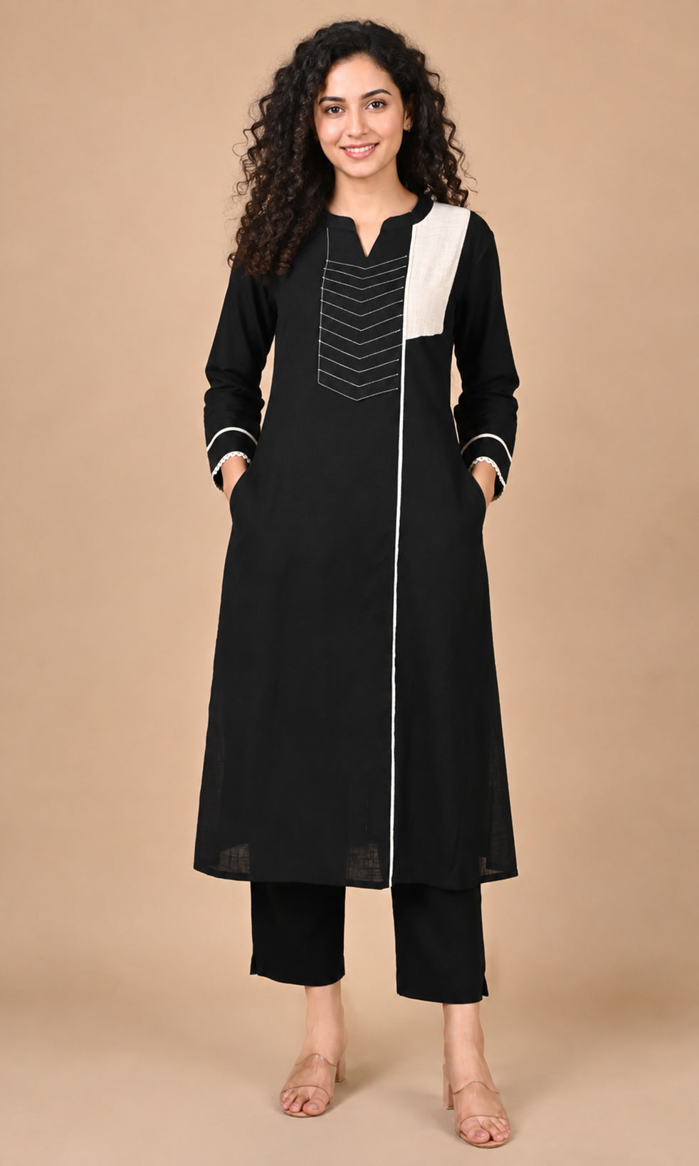 A woman modeling a black long kurta with white contrast panels and white piping details, paired with black cropped pants, against a solid beige studio background.