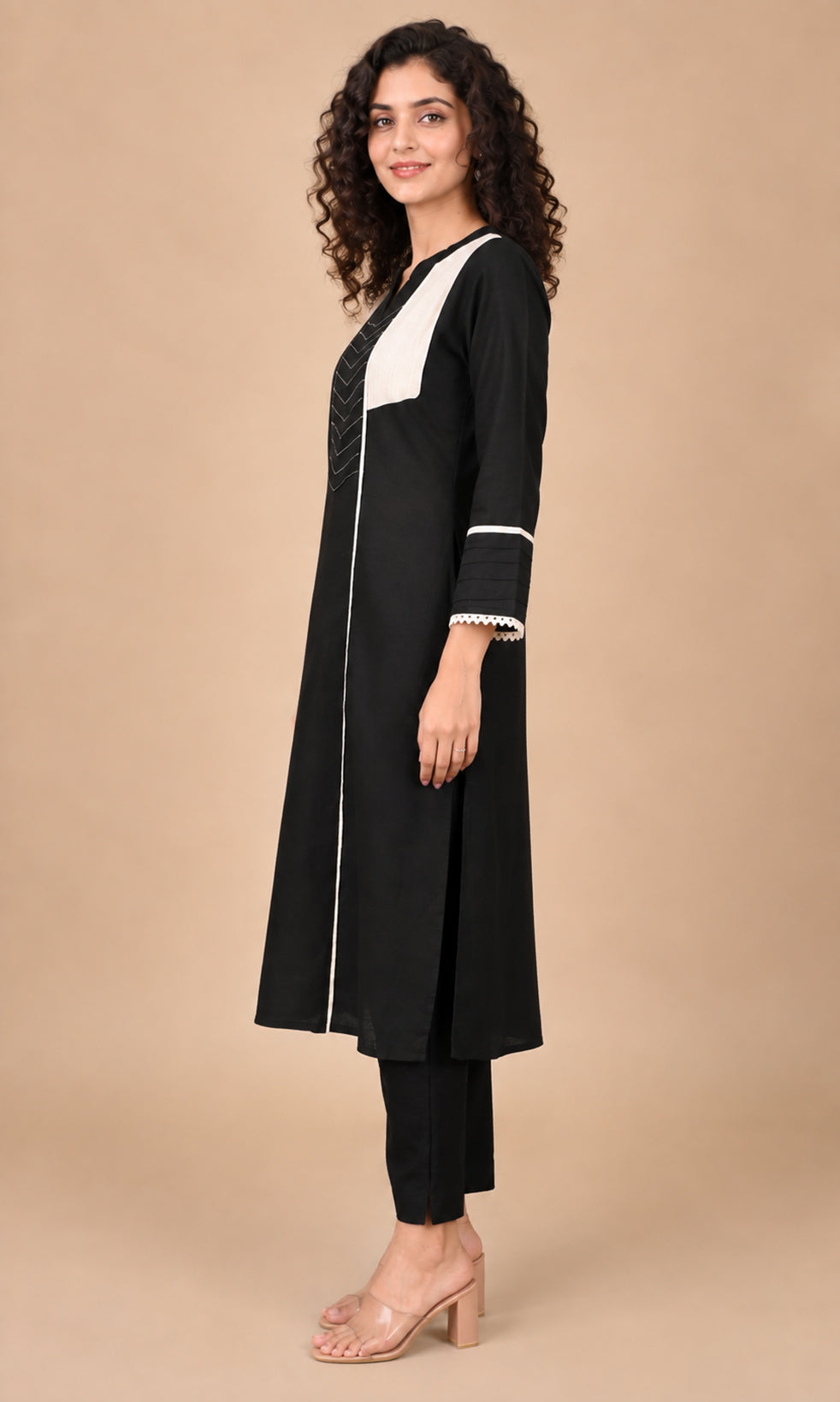 A woman modeling a black long kurta with contrasting white panels on the chest and cuffs, paired with black pants and clear high-heeled sandals, set against a solid beige studio background.