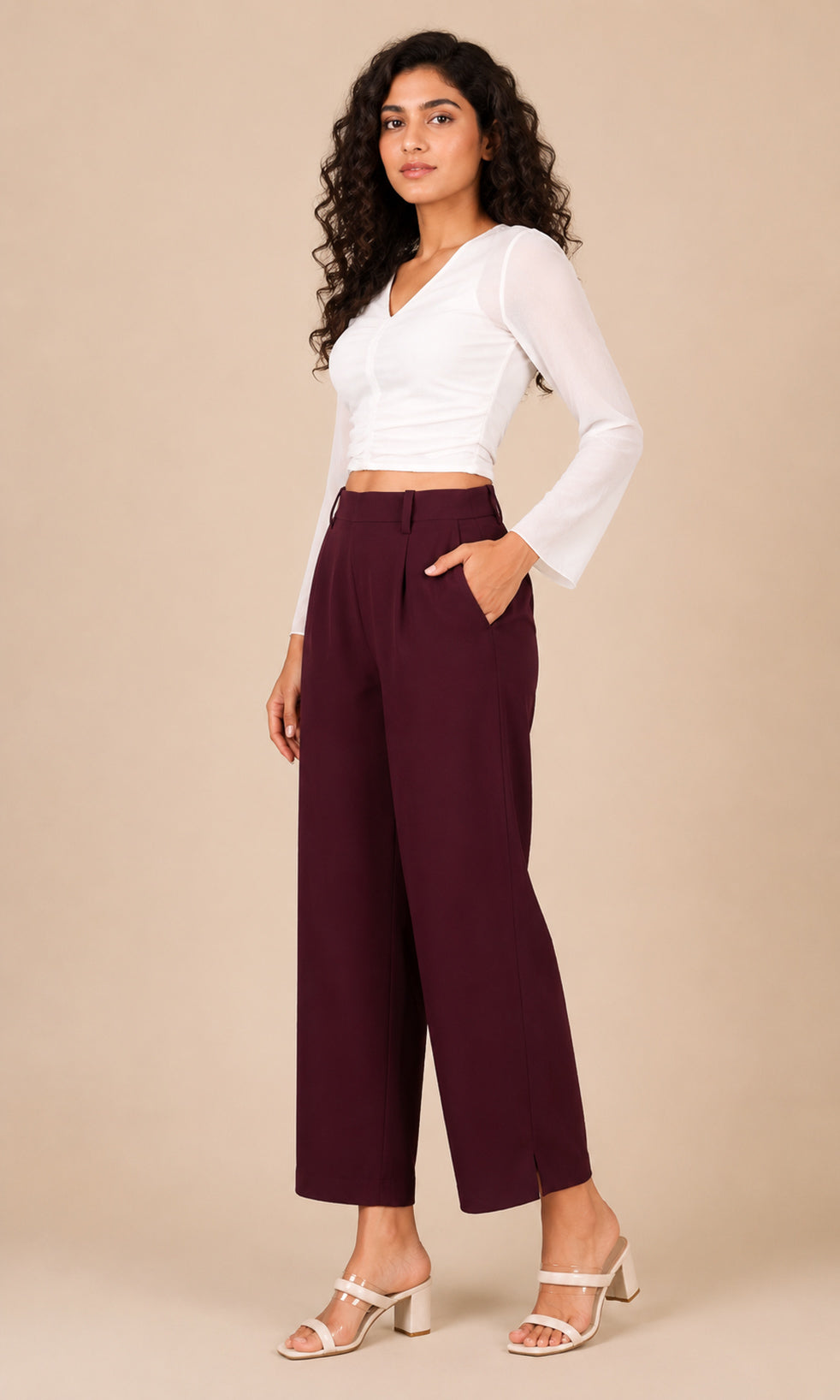 A woman modeling a white long-sleeve crop top and high-waisted burgundy wide-leg cropped pants, paired with white heeled sandals, against a solid beige studio background.