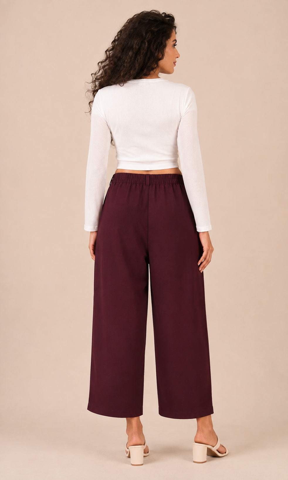 A woman shown from the back wearing a white long-sleeve crop top and dark burgundy wide-leg cropped pants, paired with white block-heel mule sandals, against a solid beige studio background.
