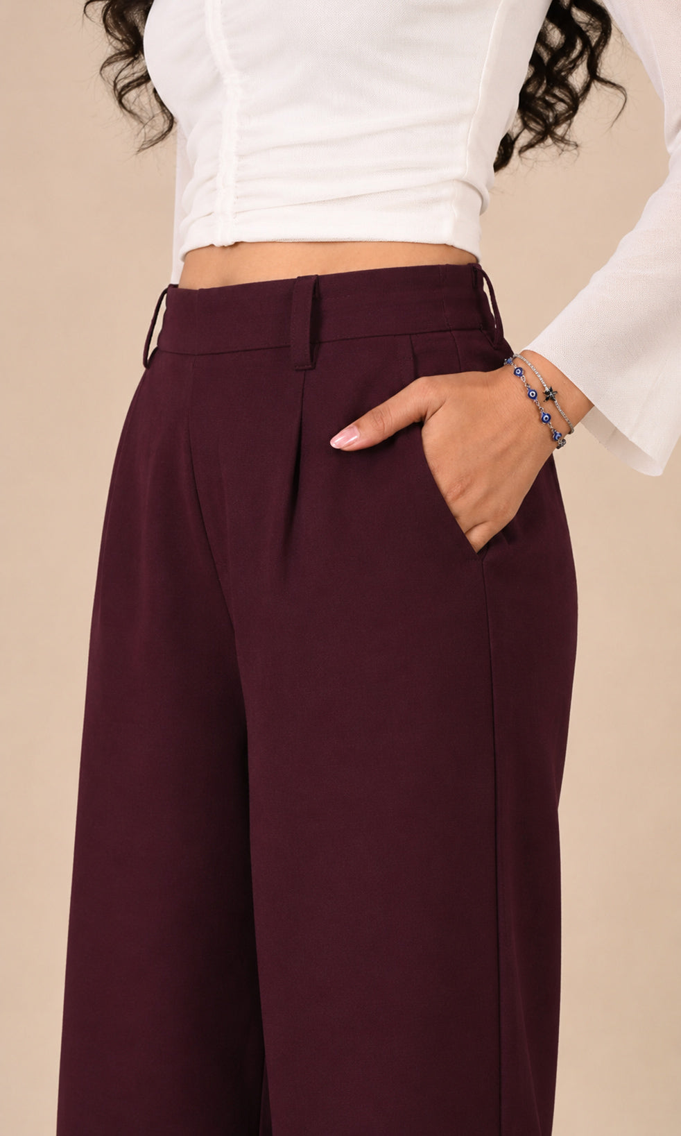 A close-up of a person wearing mid-rise, wide-leg trousers in a deep burgundy color, paired with a white cropped long-sleeve top. The person is standing against a solid beige background, with one hand in the pocket of the trousers.