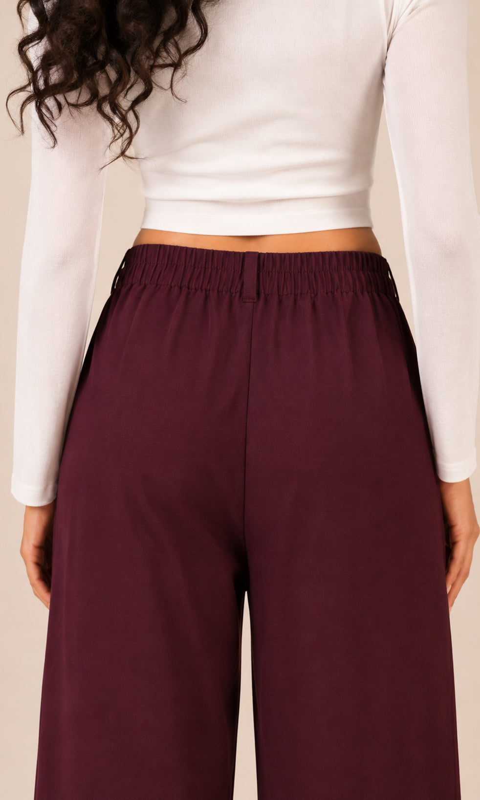 A person wearing high-waisted, wide-leg trousers in a deep burgundy or plum shade, paired with a white long-sleeve crop top. The photo is cropped from the chest down against a plain beige studio background.