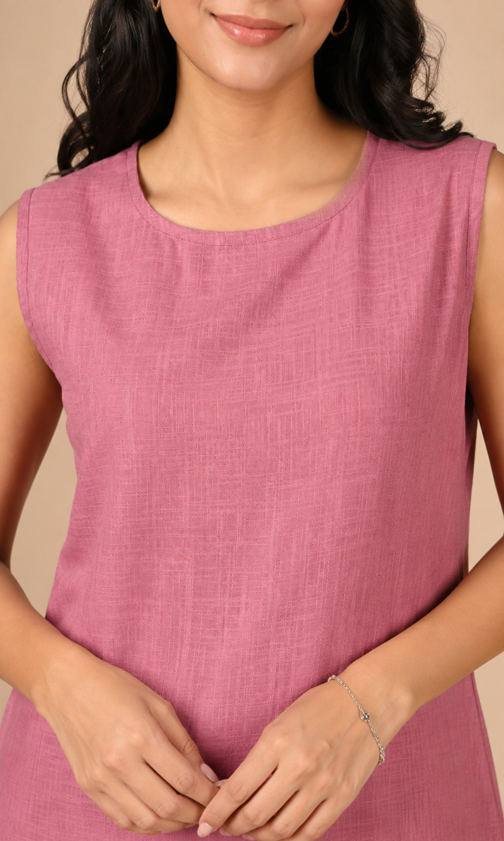 A woman wearing a sleeveless dusty pink cotton top with a round neckline, shown from the chest up against a warm beige background.
