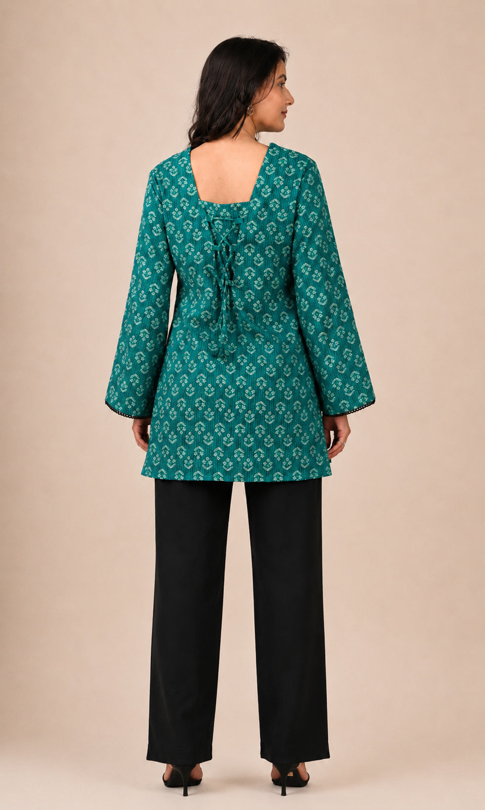 A woman shown from the back wearing a teal green patterned tunic top with black wide-leg pants, standing against a solid beige studio background.