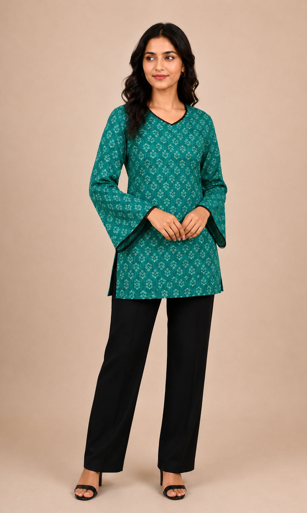 A woman modeling a teal green printed kurta with bell sleeves and black wide-leg trousers, standing against a solid beige studio background.