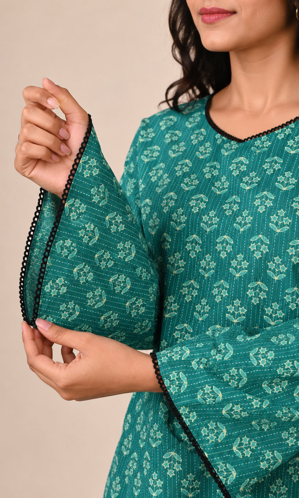 A close-up of a woman wearing a teal green printed kurta with white floral motifs and black trim on the sleeve cuffs, holding up the sleeve to show the pattern and trim.