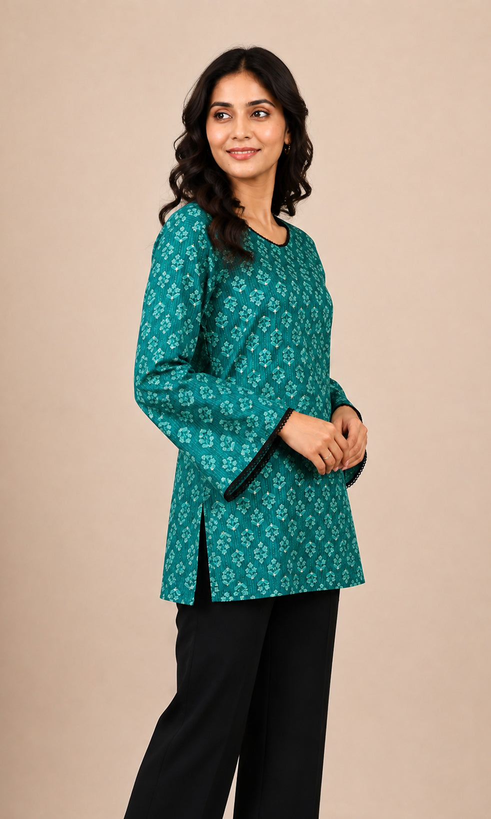 A woman modeling a teal green long-sleeve kurti with a white floral block print pattern and black bell sleeves, paired with black trousers, against a solid beige studio background.