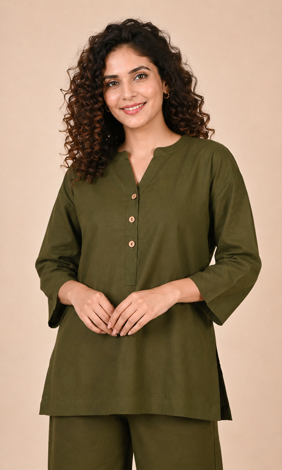 A woman with curly brown hair wearing an olive green long-sleeve Co-ord set with a buttoned split neckline, paired with matching olive green trousers, set against a solid beige background.
