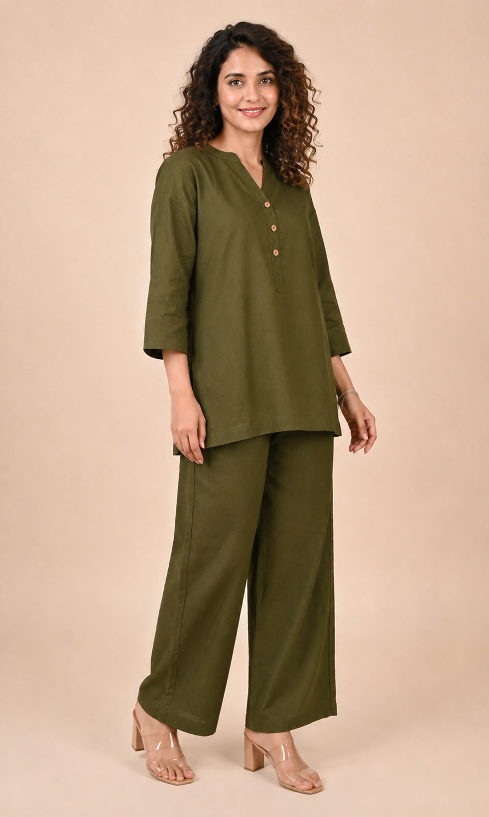 A woman modeling an olive green Co-ord set consisting of a short-sleeve buttoned Co-ord top and matching wide-leg trousers, paired with clear high-heeled sandals, against a solid beige studio background.