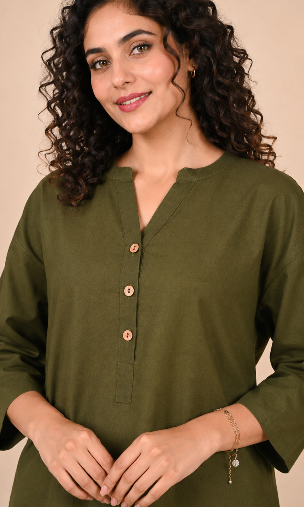 A woman with curly dark hair wearing an olive green long-sleeveCo-ord set top with a split neckline and three decorative buttons, posed against a solid beige background.