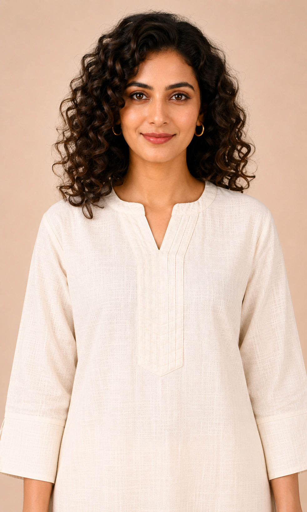 A woman with curly dark hair wearing a cream-colored linen tunic top with a split neckline, photographed against a solid beige background.