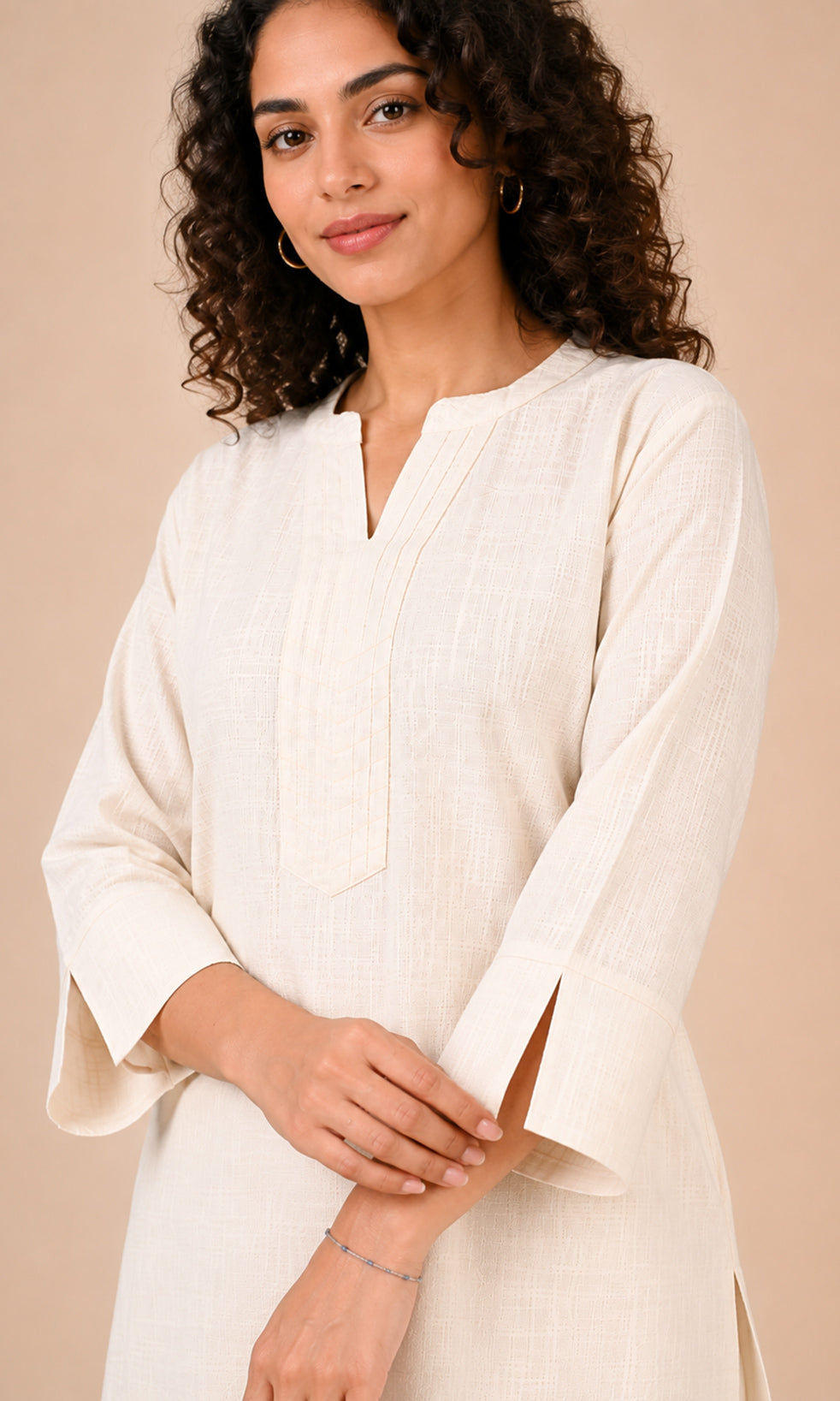 A woman with curly dark hair wearing a cream-colored cotton a-line kurta with a split neckline and wide three-quarter sleeves, posed against a solid beige background.