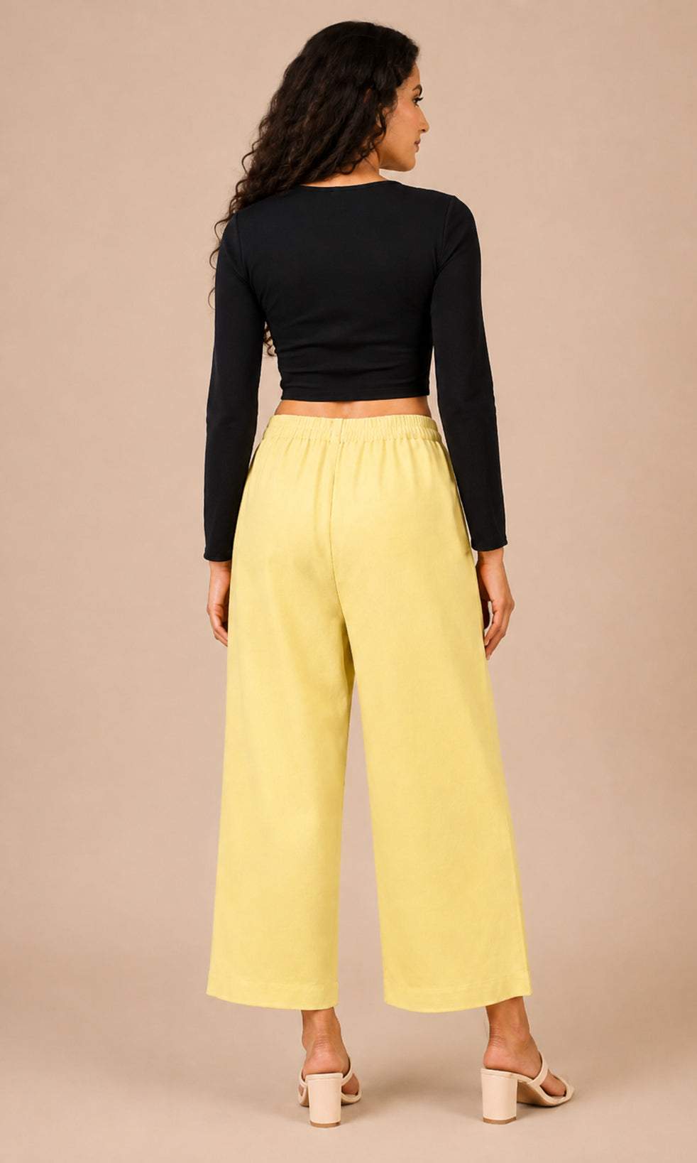 A woman shown from the back wearing a black long-sleeve crop top and light yellow wide-leg pants, standing against a solid beige studio background.
