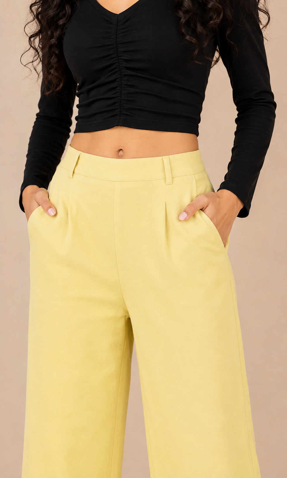 A model wearing high-waisted, wide-leg yellow trousers with pleated details and side pockets, paired with a black ruched long-sleeve crop top, set against a solid beige background.