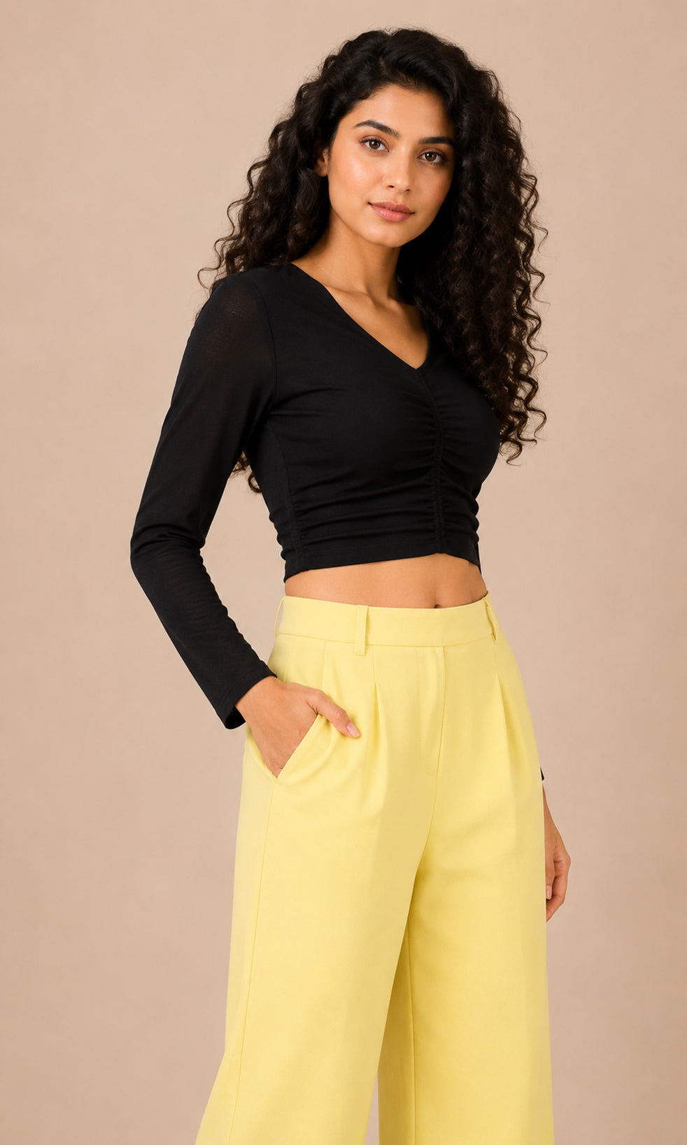 A woman modeling a black long-sleeve ruched crop top paired with light yellow high-waisted wide-leg pants, set against a solid beige studio background.