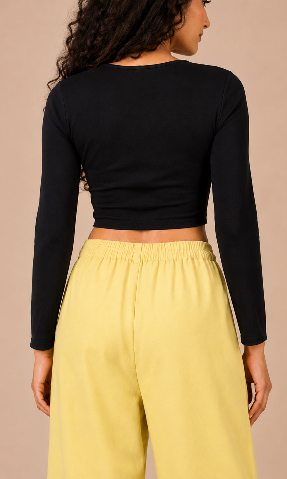 A woman shown from the back wearing a black long-sleeve cropped top and light yellow wide-leg pants against a solid beige background.