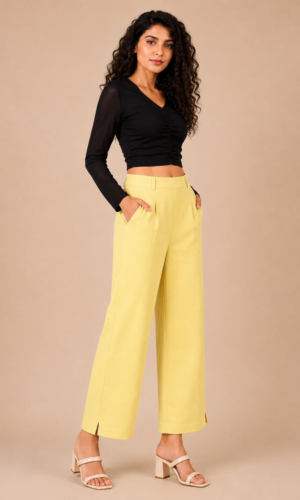A woman modeling a black long-sleeve crop top and light yellow wide-leg cropped pants with side slits, paired with beige heeled sandals, against a solid beige studio background.