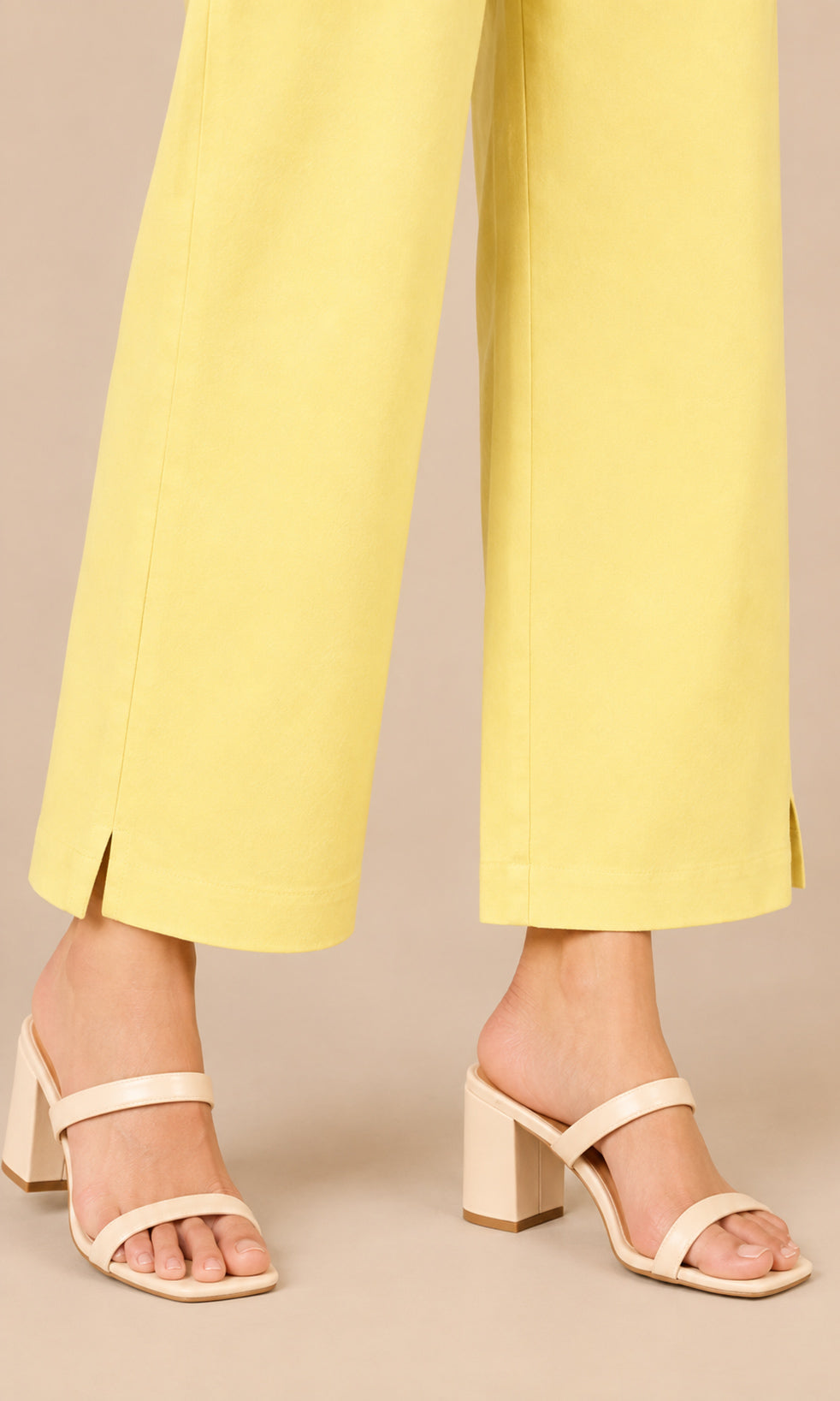 A close-up of a person's feet wearing pale yellow wide-leg cropped pants and light beige block-heel slide sandals against a solid beige studio background.