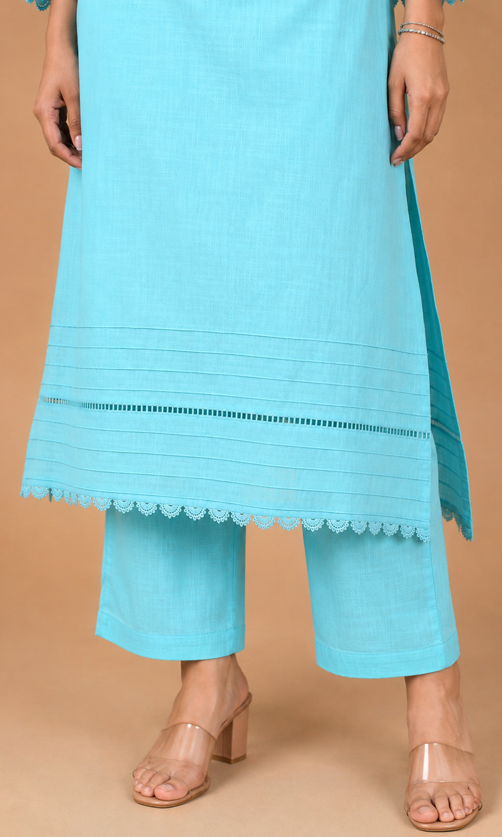 A model wearing a light blue kurta and matching cropped pants with scalloped lace trim, paired with clear heeled sandals, set against a solid beige background.