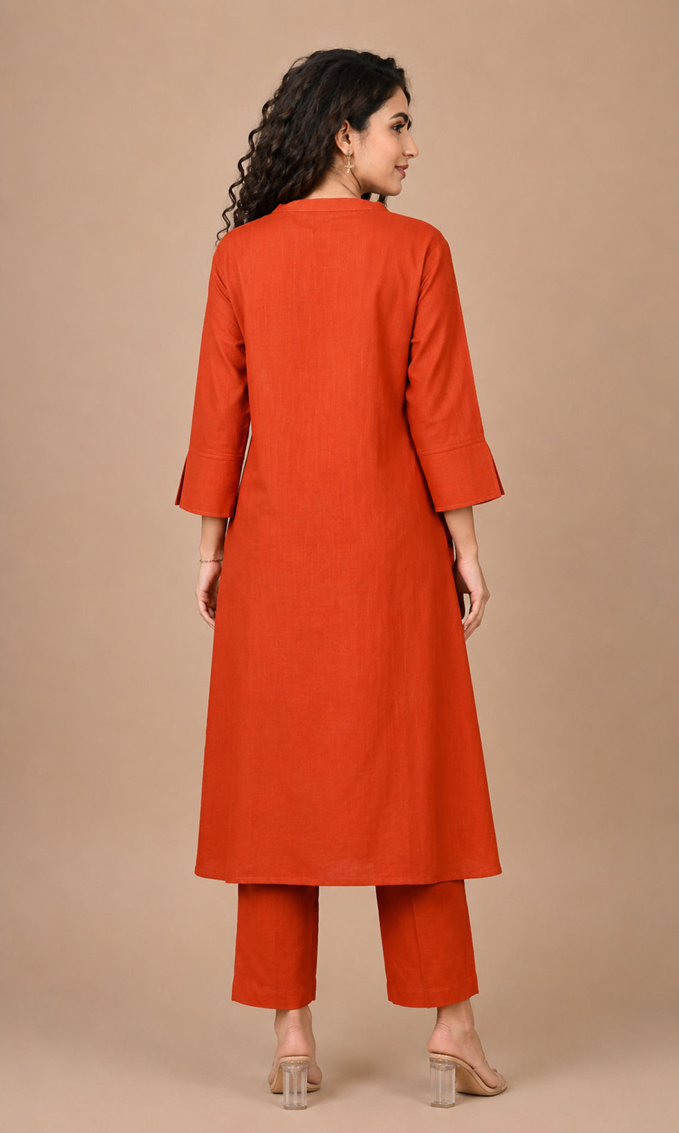 A woman is shown from the back wearing a bright rust-orange kurta and matching pants set against a solid beige studio background.