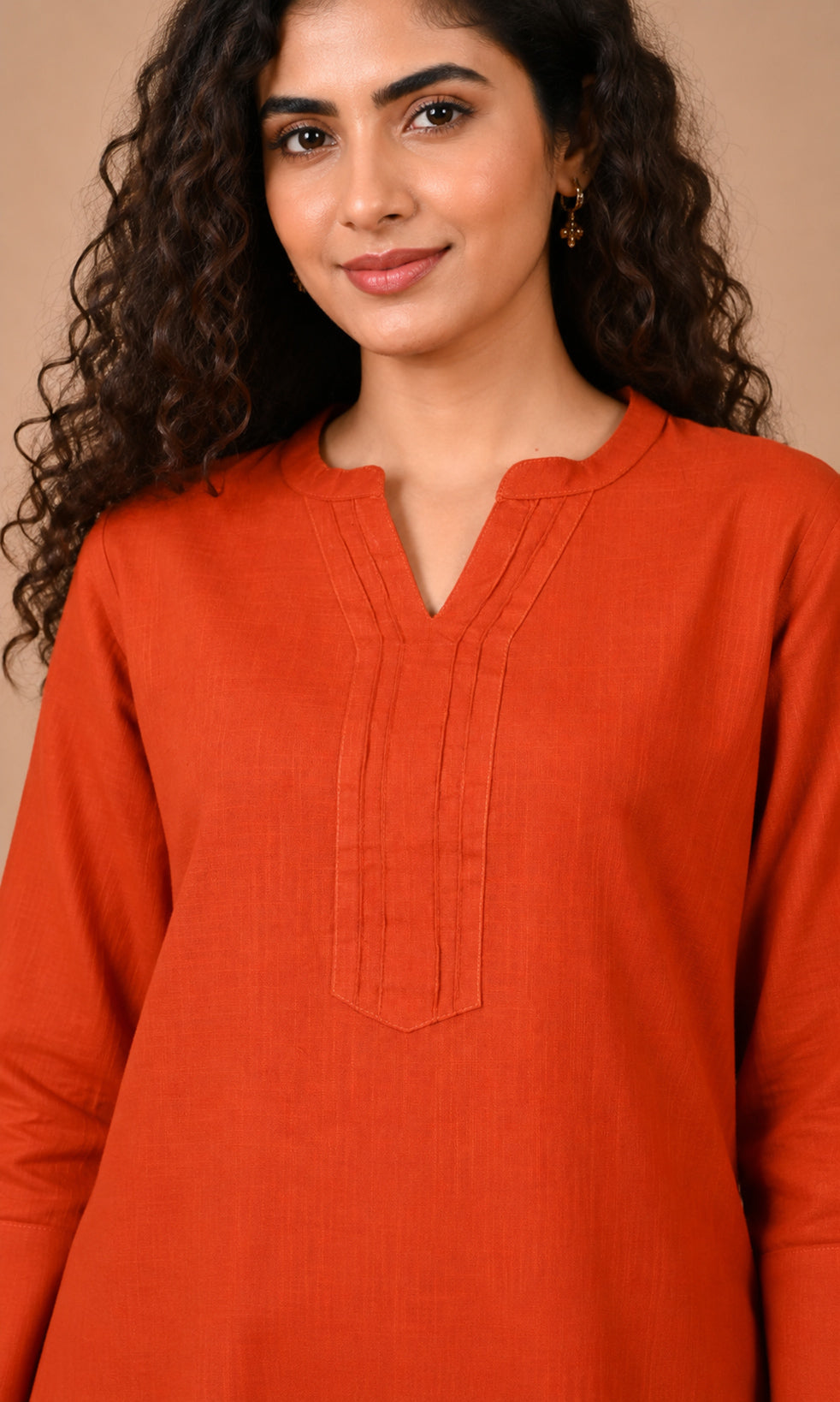 A woman with long, curly dark brown hair wearing a rust-orange long-sleeve kurta with a split V-neck and vertical pleat detailing on the chest, set against a solid warm beige background.