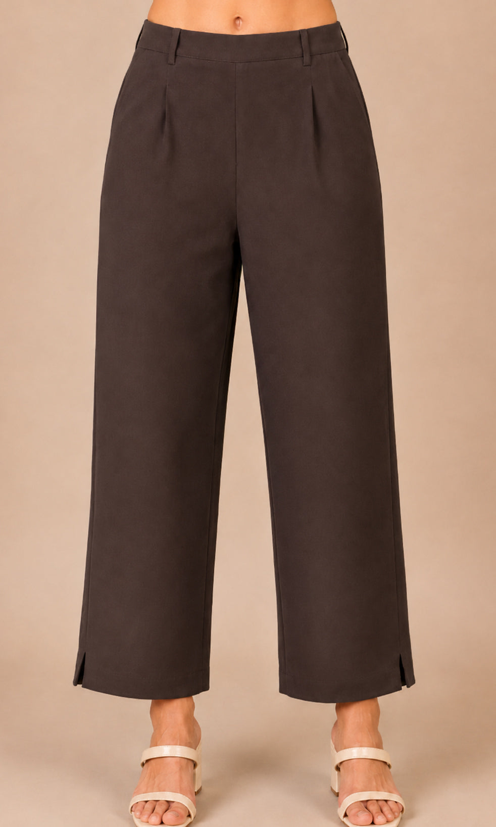 A person wearing dark brown cropped wide-leg pants with side slits and pleats, paired with white heeled sandals, against a plain beige studio background.