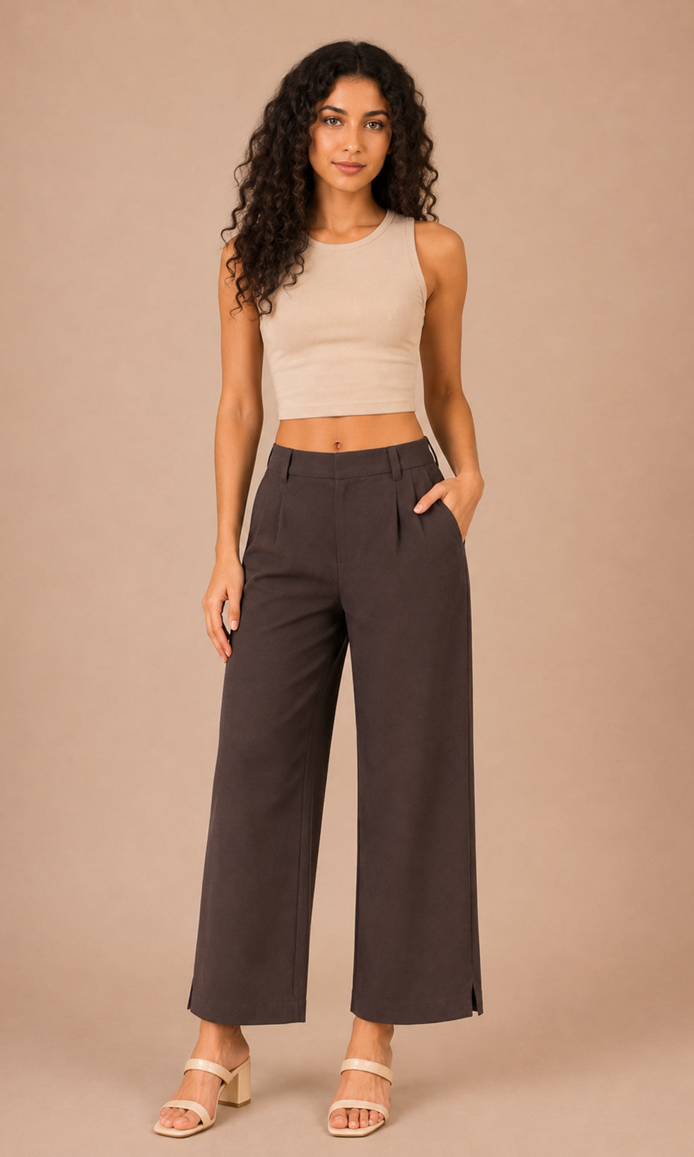 A woman modeling a pair of dark brown wide-leg cropped pants with a beige ribbed crop top, standing against a solid beige studio background.