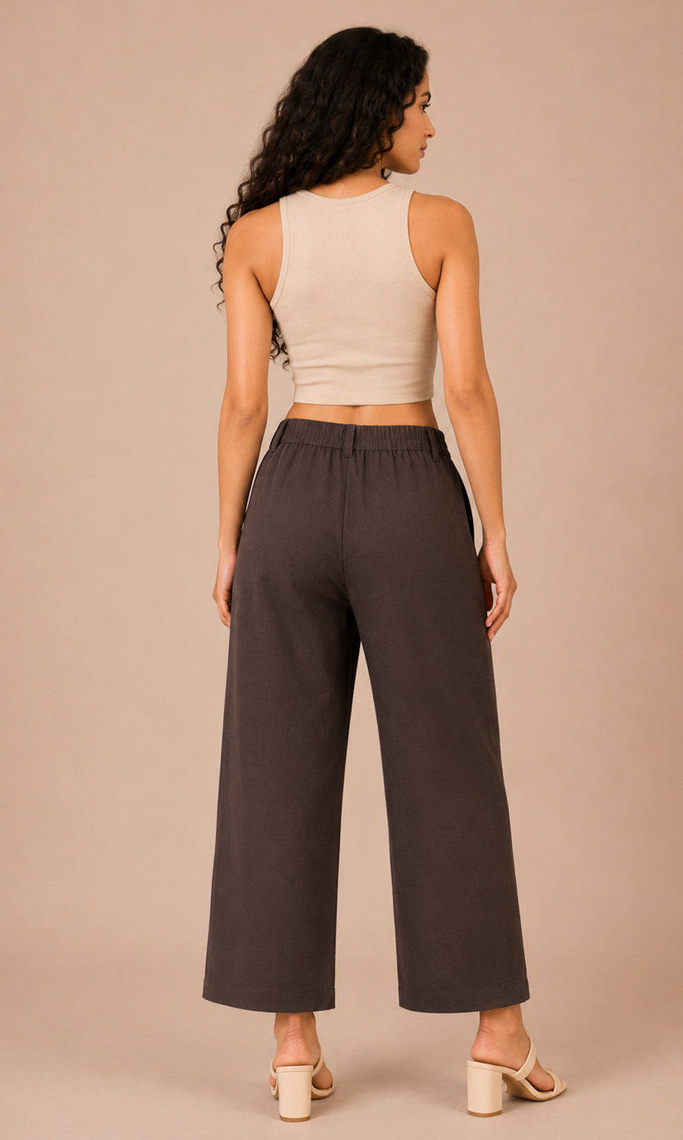 A woman shown from the back wearing dark charcoal wide-leg cropped pants and a light beige sleeveless crop top, standing against a solid beige studio background.