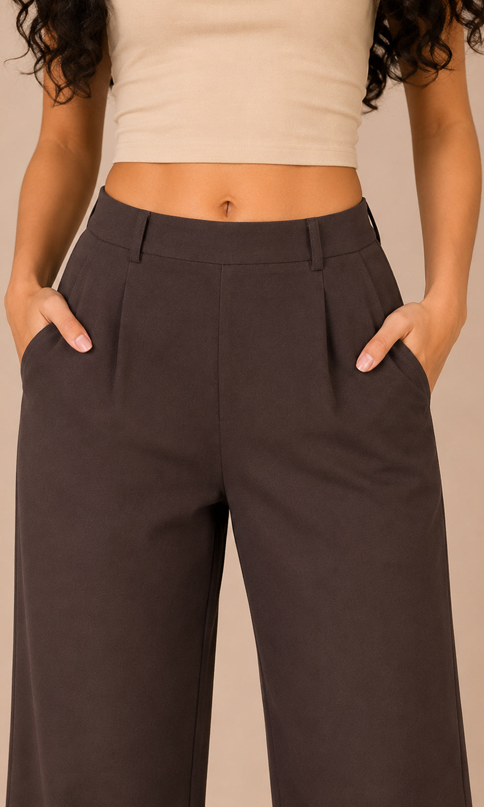 A close-up of a model wearing dark brown wide-leg trousers with pleated details and side pockets, paired with a beige crop top against a solid beige background.