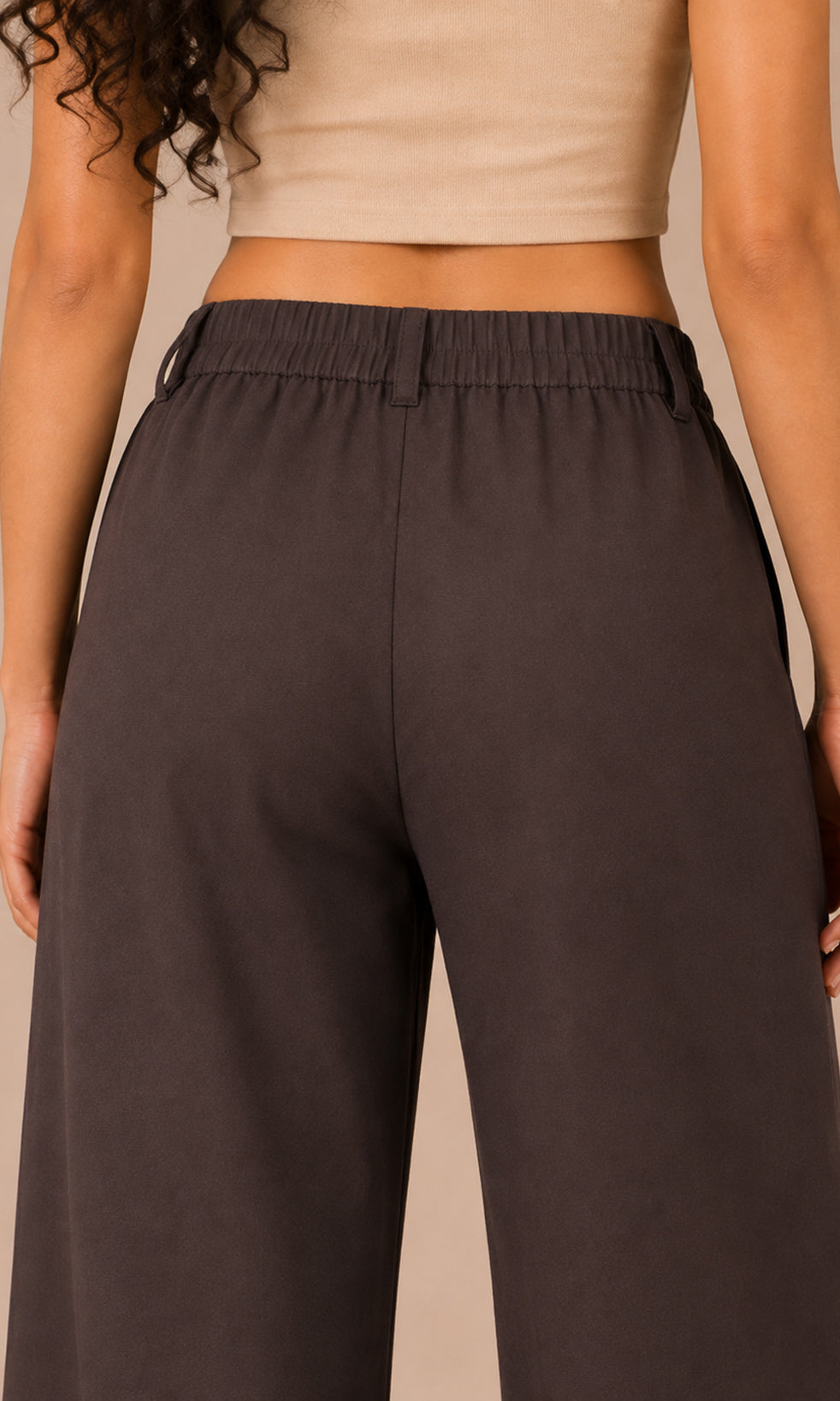 A close-up back view of a model wearing dark brown wide-leg pants with an elastic waistband, paired with a beige crop top against a solid beige background.