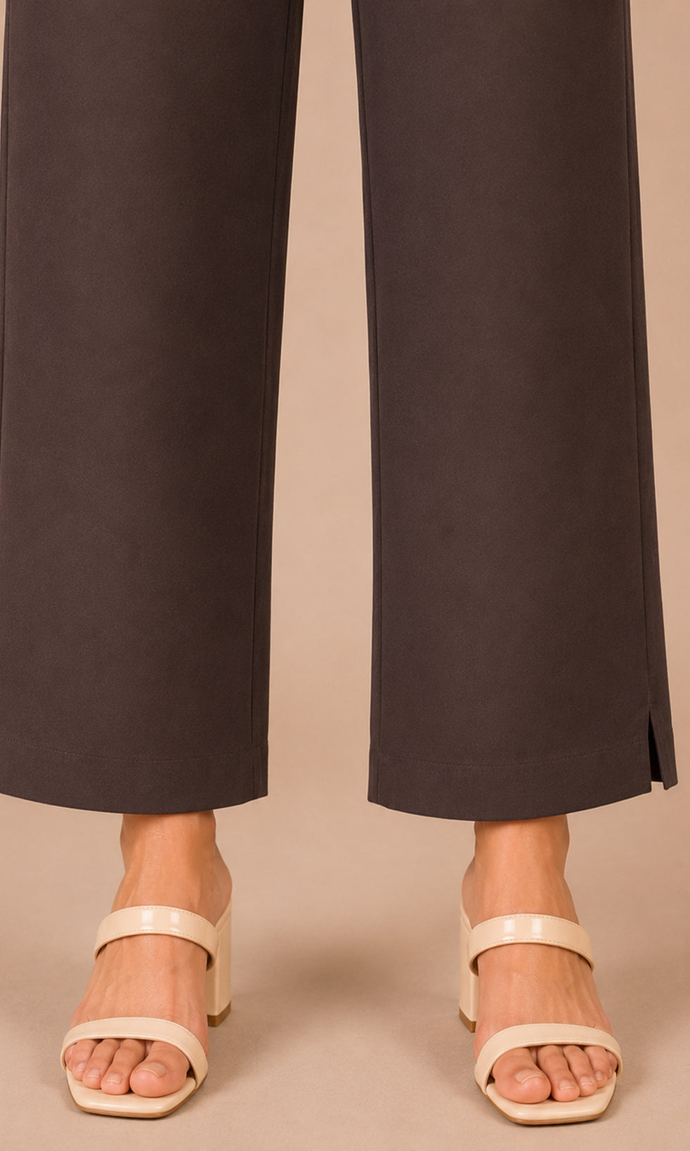 A close-up of a person's lower legs wearing dark brown wide-leg trousers and light beige block-heel sandals against a solid beige background.