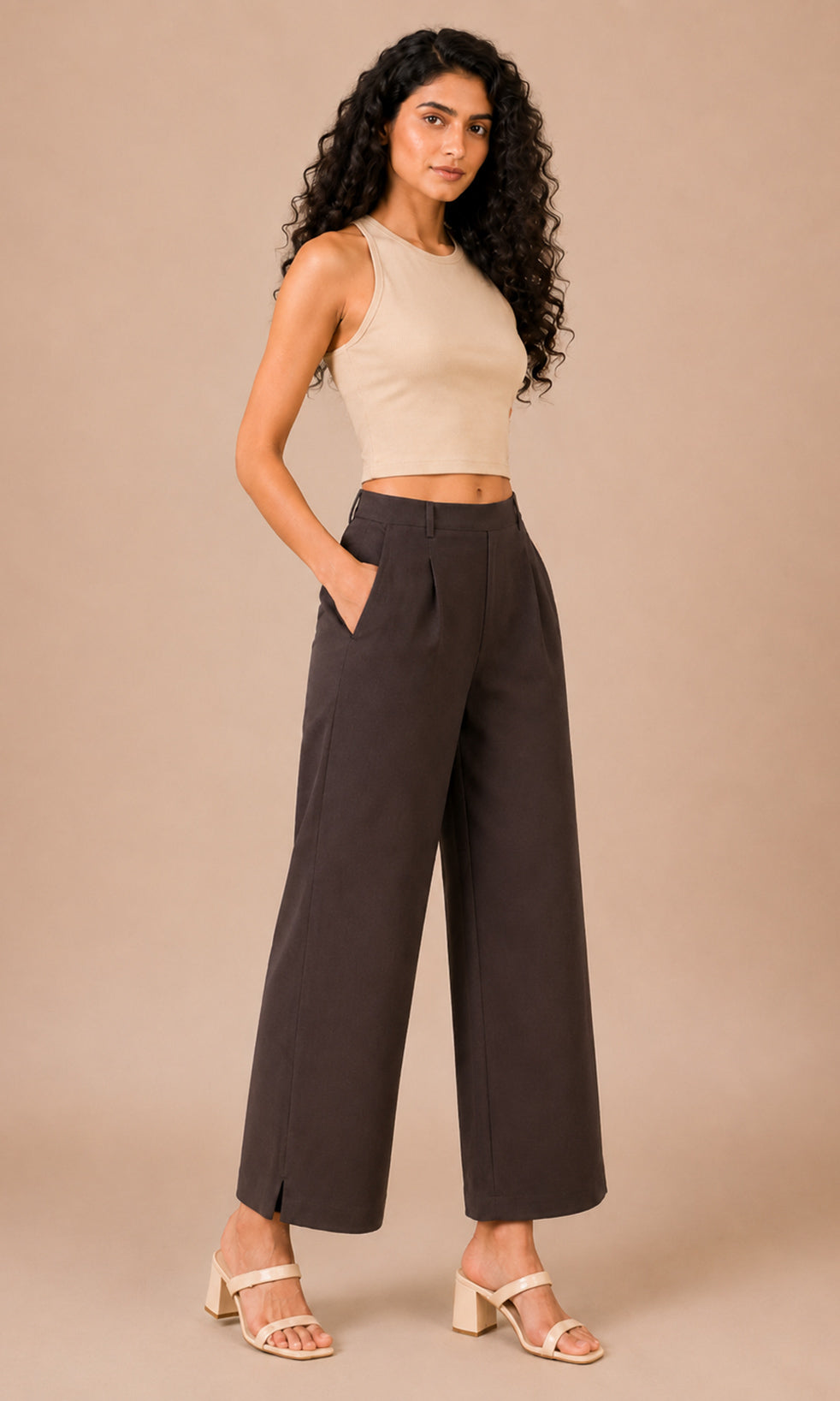 A woman modeling a pair of dark brown wide-leg cropped pants with a beige sleeveless crop top, standing against a solid beige studio background.